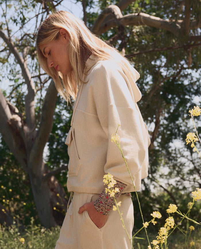 A person wearing THE GREAT. SP26 TGO The Basecamp Pullover in Oatmilk stands outdoors among yellow wildflowers and trees, looking down.