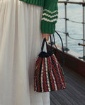 A person in a green sweater and white skirt holds THE GREAT. SU26 The Beaded Cinched Bucket Bag – Cabana Stripe while standing on a wooden deck near the water.
