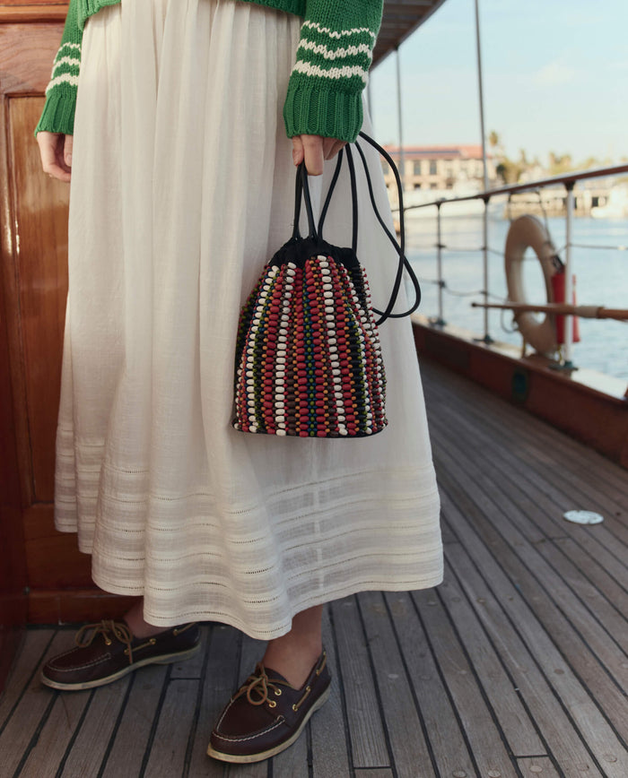 A person holds THE GREAT. SU26's The Beaded Cinched Bucket Bag in Cabana Stripe while standing on a wooden deck, dressed in a white skirt, green sweater, and brown boat shoes.