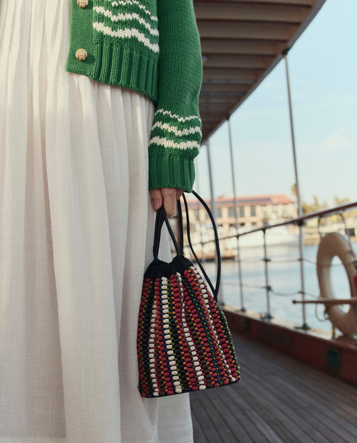 A person in a green sweater and white skirt stands on a dock, holding THE GREAT. SU26 The Beaded Cinched Bucket Bag – Cabana Stripe.