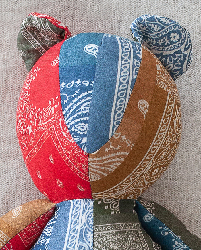 The Bear. -- Multi by THE GREAT. features a patchwork cotton design in red, blue, brown, and green prints, capturing the nostalgic charm of an heirloom toy against a light textured backdrop.