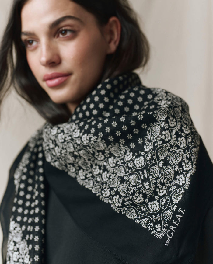 A woman wearing THE GREAT. CARRYOVER's "The Big Bandana. -- Almost Black," an oversized black shawl with white floral patterns and "THE GREAT." text by the corner, looks off to the side.