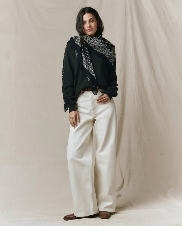 A woman stands against a beige backdrop wearing THE GREAT. CARRYOVER's The Big Bandana in Almost Black, styled with a black top, white wide-leg pants, brown shoes, and a brown belt.