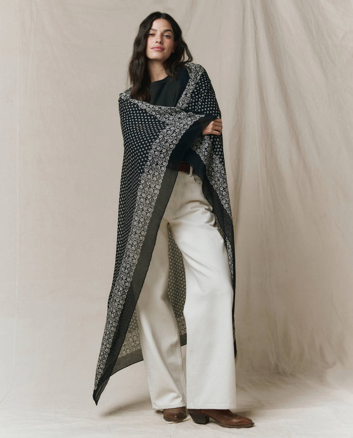 A woman stands before a beige backdrop wearing THE GREAT. CARRYOVER’s The Big Bandana in Almost Black over a black top, white wide-leg pants, and brown shoes.