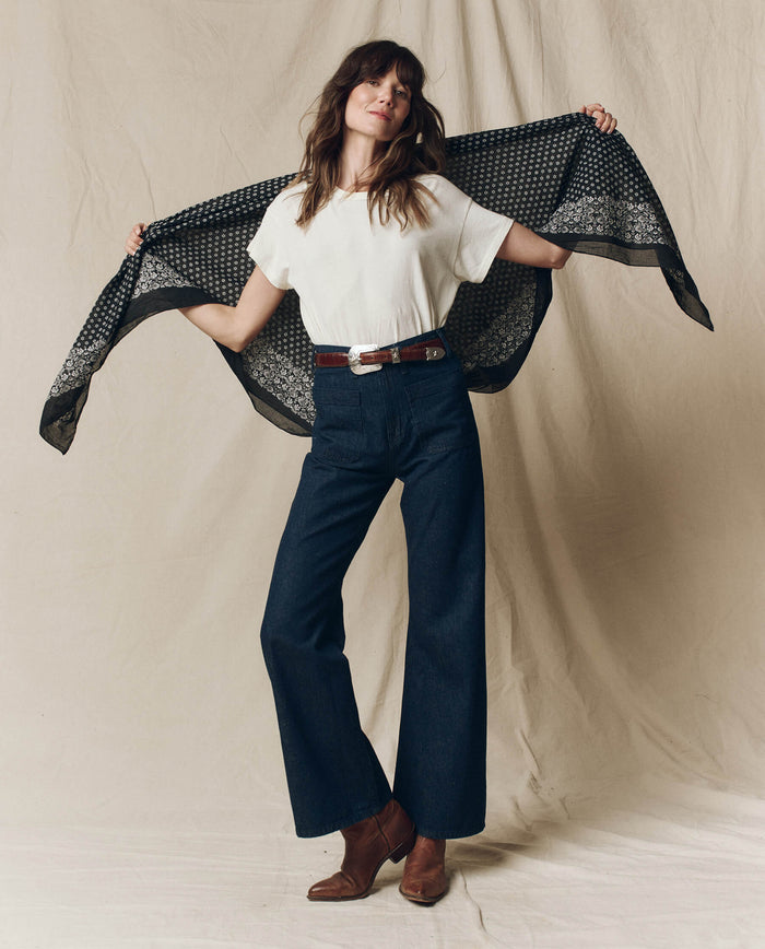 A person poses with The Big Bandana in Almost Black by THE GREAT. SP25 D2 spread behind them, wearing a white t-shirt, dark flared jeans, and brown boots against a neutral background.