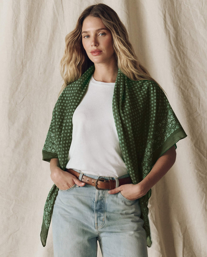 A woman with long wavy hair wears a white T-shirt, light blue jeans, and a brown belt, with THE GREAT. F26 BIG BANDANA in Army draped over her shoulders, standing against a neutral fabric backdrop.