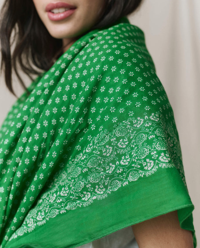 A woman wears THE GREAT. SP26 D1 "The Big Bandana – Bright Sprig," a green shawl with white floral patterns and a decorative border, draped over her shoulder in a vintage bandana style.