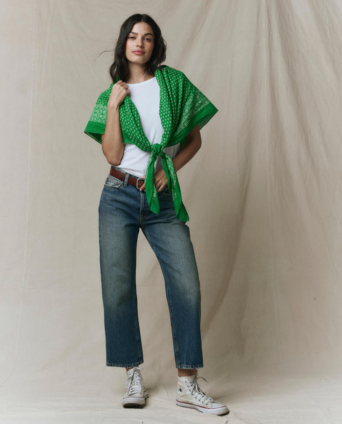 A person stands against a neutral backdrop wearing a white tee, blue jeans, white sneakers, and THE GREAT. SP26 D1 The Big Bandana in Bright Sprig tied at the front.