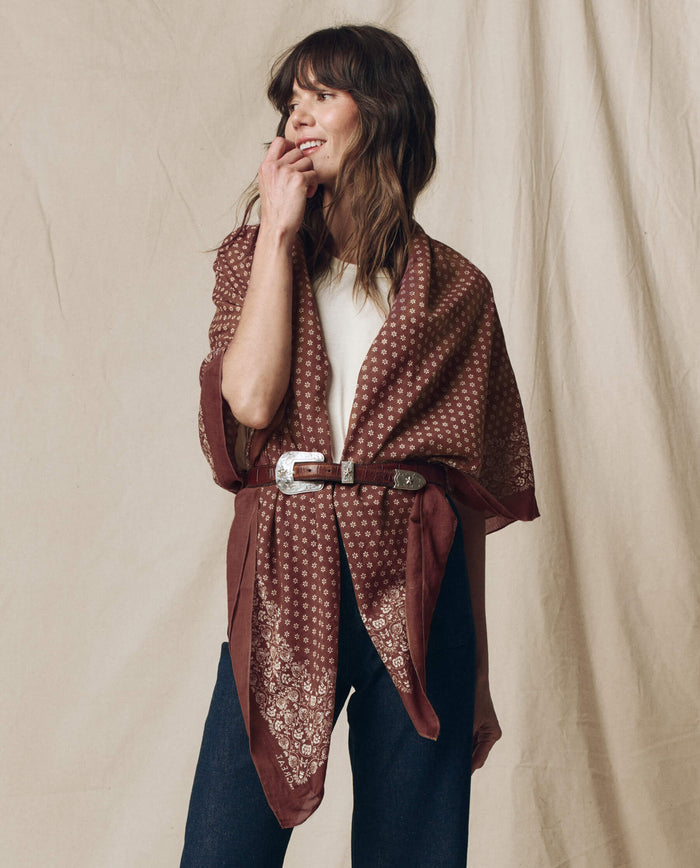 A woman wears THE GREAT. F26 BIG BANDANA in Chocolate, styled over a white top and dark jeans with a wide belt at the waist, standing before a beige fabric backdrop.