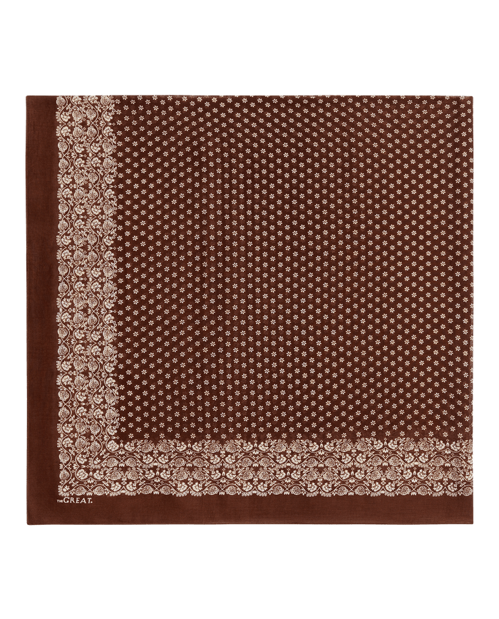 THE GREAT. F26 BIG BANDANA – The Big Bandana in Chocolate features a white floral and dot pattern on a brown square, with decorative borders along two edges reminiscent of a vintage handkerchief.