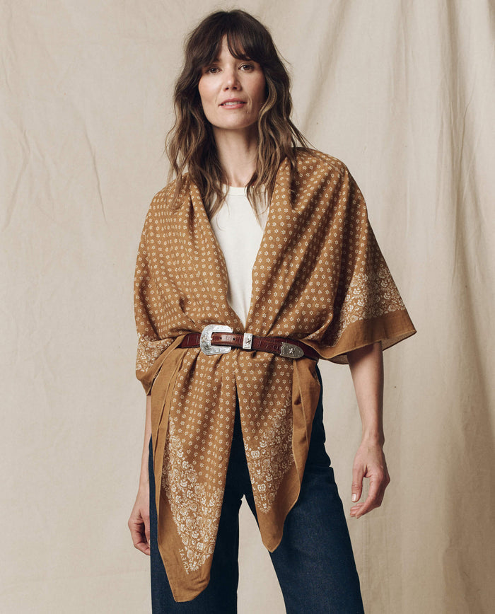 A person elegantly stands against a beige background wearing THE GREAT. SP25 D2's The Big Bandana in Marigold, belted over a white shirt with dark jeans.