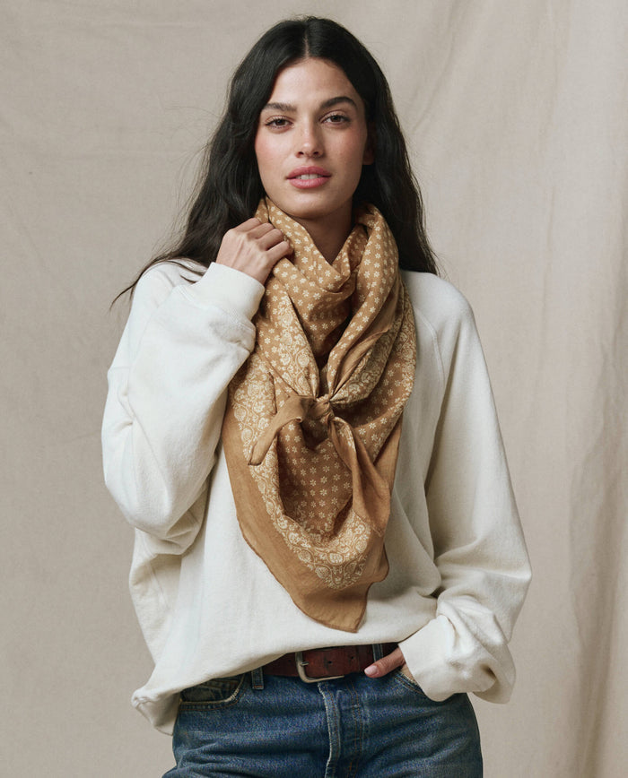 A person with long dark hair wearing a white sweater, blue jeans, and The Big Bandana in Marigold by THE GREAT. CARRYOVER stands against a neutral fabric backdrop.