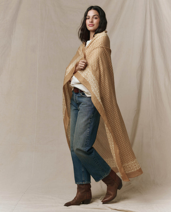 A woman stands on a beige background wearing blue jeans, a white shirt, brown boots, and The Big Bandana in Marigold by THE GREAT. CARRYOVER draped over her shoulders.