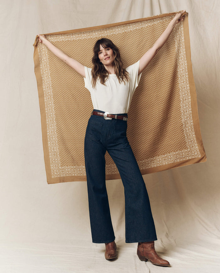 On a neutral backdrop, a person in a white shirt and jeans displays The Big Bandana in Marigold by THE GREAT. SP25 D2, made of 100% cotton.