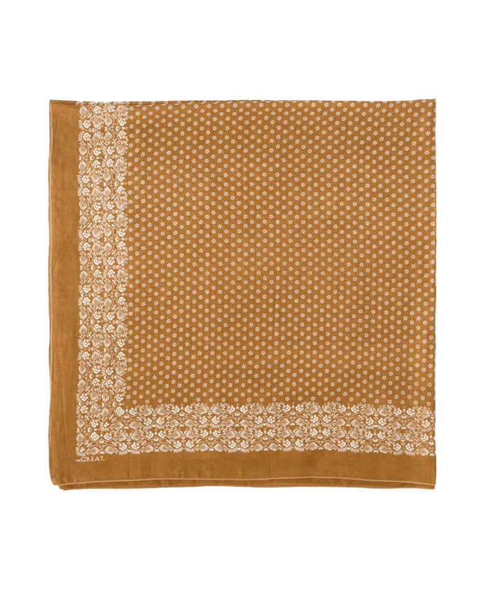 The Big Bandana in Marigold by THE GREAT. CARRYOVER is a square brown cotton scarf featuring a white floral border and delicate white floral patterns—a charming vintage accessory for any occasion.
