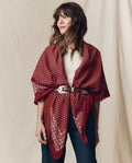 A woman wearing THE GREAT. F26 BIG BANDANA in maroon, styled as a shawl belted at the waist over a white top and jeans, standing in front of a beige backdrop.