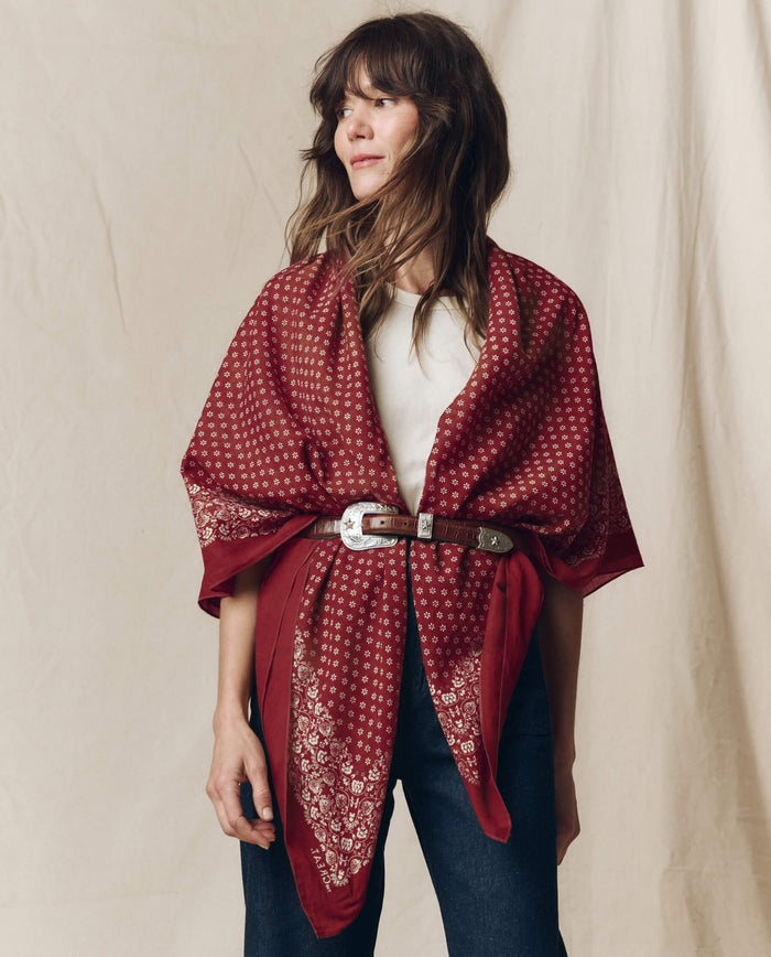 A woman wearing THE GREAT. F26 BIG BANDANA in maroon, styled as a shawl belted at the waist over a white top and jeans, standing in front of a beige backdrop.