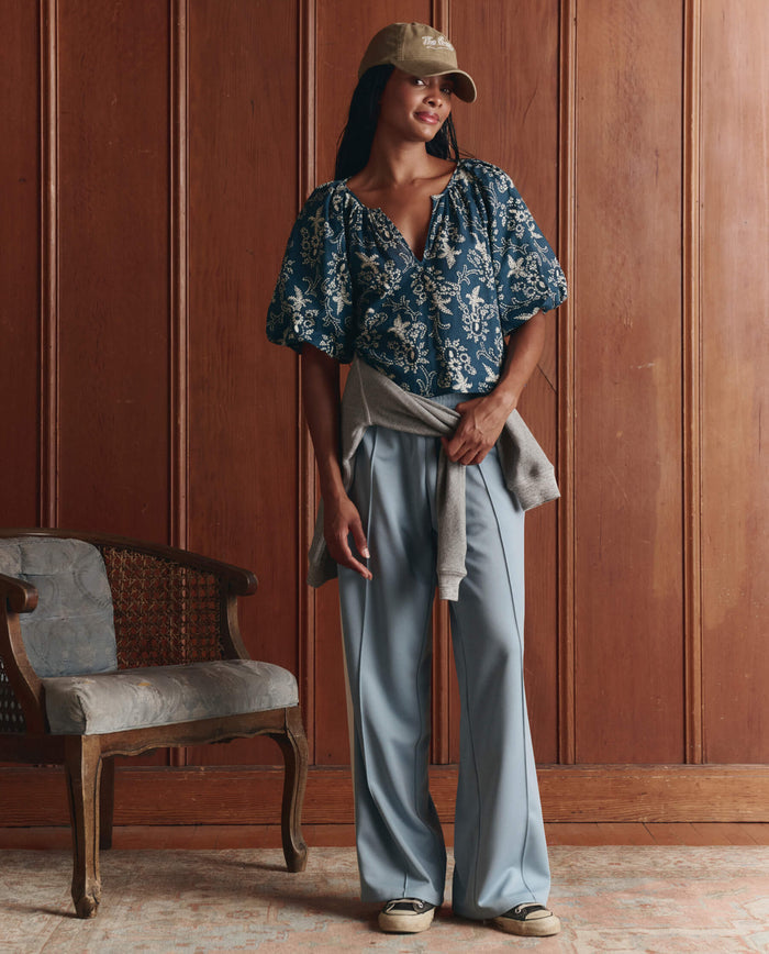 A woman stands indoors in front of a wooden paneled wall, wearing THE GREAT. SP26 D1 The Blythe Top. -- Indigo Bandana, light blue trousers, sneakers, a beige cap, and a gray sweater tied around her waist.
