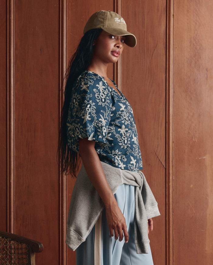 A woman in THE GREAT. SP26 D1 The Blythe Top – Indigo Bandana, blue pants, and a tan cap stands sideways by a wooden wall, with a gray sweatshirt tied around her waist.
