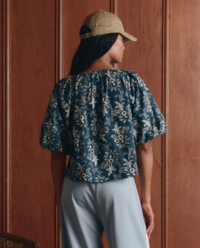A person wearing a khaki cap, THE GREAT. SP26 D1 The Blythe Top -- Indigo Bandana with puff sleeves, and light blue pants stands indoors facing a wooden paneled wall.