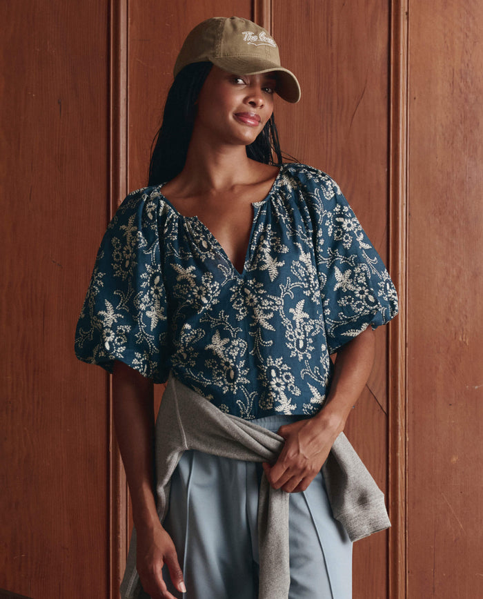 A person in THE GREAT. SP26 D1's The Blythe Top—Indigo Bandana, light blue pants, a tan baseball cap, and a gray sweater tied at the waist stands before a wooden wall.
