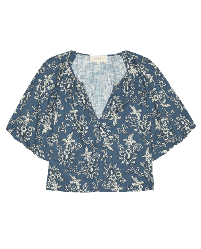 The Blythe Top by THE GREAT. SP26 D1 is a chic indigo bandana short-sleeve women’s blouse with a white floral pattern, v-neckline, and slightly cropped hem.