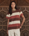 A person with long dark hair, dressed in THE GREAT. PF26 The Boxy Crew. -- Ashwood Stripe and light pants, stands before an ornately carved wooden door.