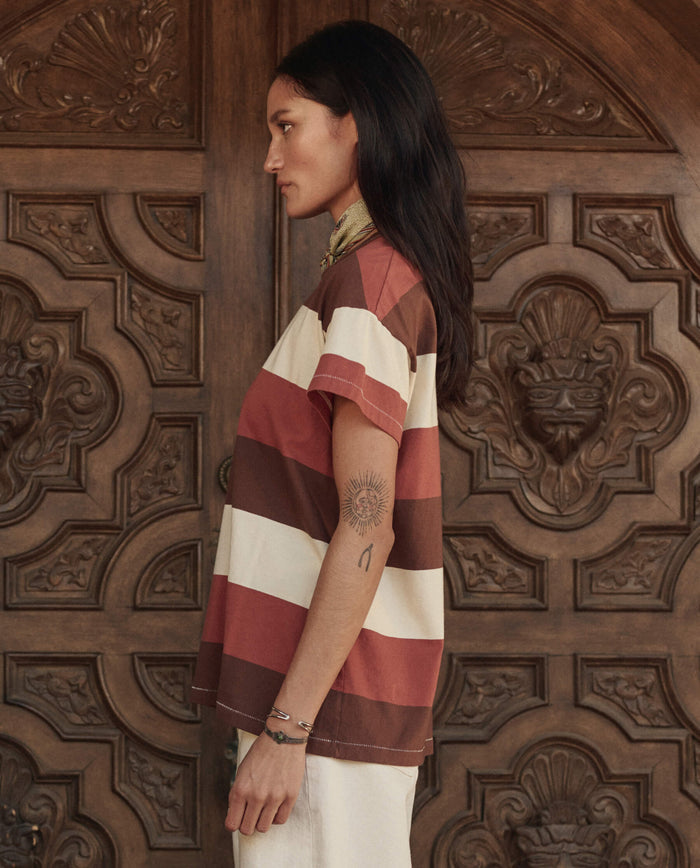 Woman with long dark hair in profile, wearing THE GREAT. PF26 The Boxy Crew. -- Ashwood Stripe in brown and cream stripes, stands before an ornate wooden door, her left arm tattoos showing off a handcrafted Los Angeles style.