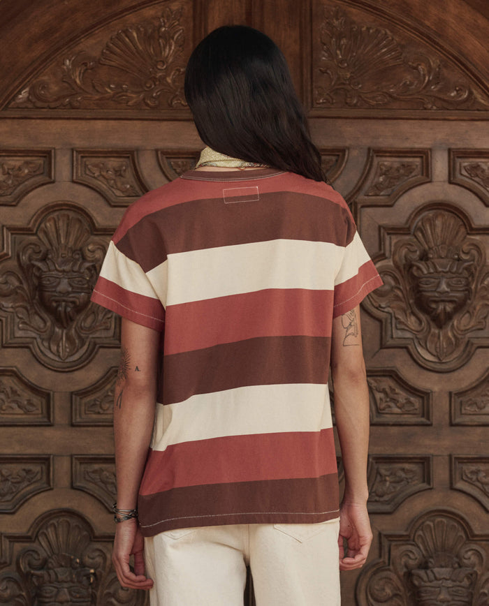 Person with long hair, back to camera, wears THE GREAT. PF26 The Boxy Crew. -- Ashwood Stripe tee and light pants, standing before an ornately carved handcrafted Los Angeles wooden door.