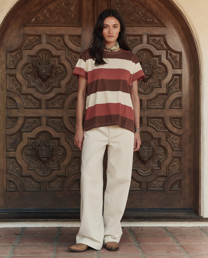 A woman stands before an ornate wooden door, wearing THE GREAT. PF26 The Boxy Crew. -- Ashwood Stripe t-shirt, wide-leg cream pants, and brown shoes.