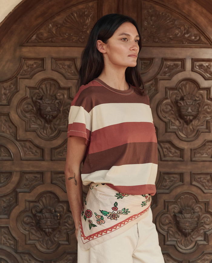 A woman with long dark hair stands before an ornate wooden door, wearing THE GREAT. PF26 The Boxy Crew. -- Ashwood Stripe and cream floral embroidered shorts.