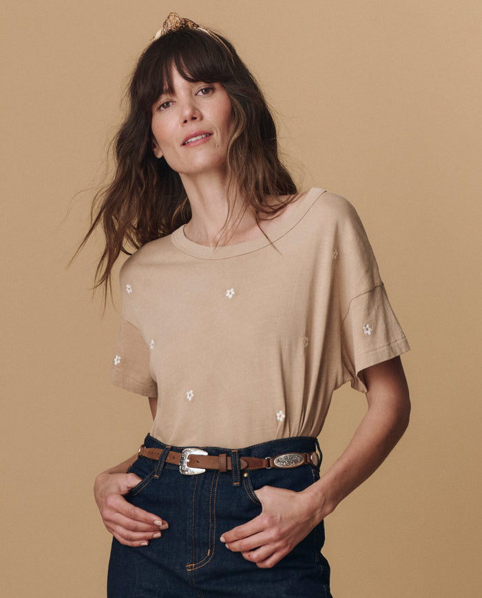 A woman wears THE GREAT. PF25 EMB KNITS The Boxy Crew. Embroidered -- Khaki with Daisy Embroidery, styled with dark high-waisted jeans and a belt, standing against a plain beige background.