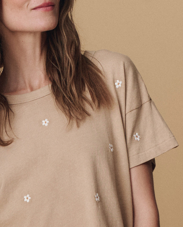 A woman wears THE GREAT. PF25 EMB KNITS 'The Boxy Crew. Embroidered' t-shirt in khaki with daisy embroidery, standing against a matching background. Only her lower face, neck, and upper torso are visible.
