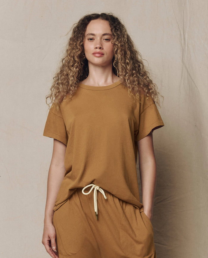 A woman with curly hair wears THE GREAT.'s The Boxy Crew. Solid in Cork, paired with matching drawstring pants, standing against a neutral background.