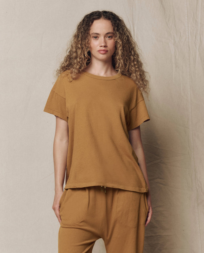 A person with long curly hair wears THE GREAT.'s The Boxy Crew. Solid in Cork, paired with matching pants, standing against a neutral fabric backdrop.