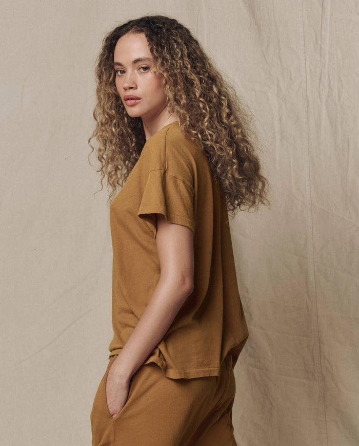 A person with long, curly hair stands sideways against a plain beige backdrop, wearing THE GREAT. SU26 SEASONAL CORE's The Boxy Crew. Solid -- Cork t-shirt and pants, with one hand in their pocket.