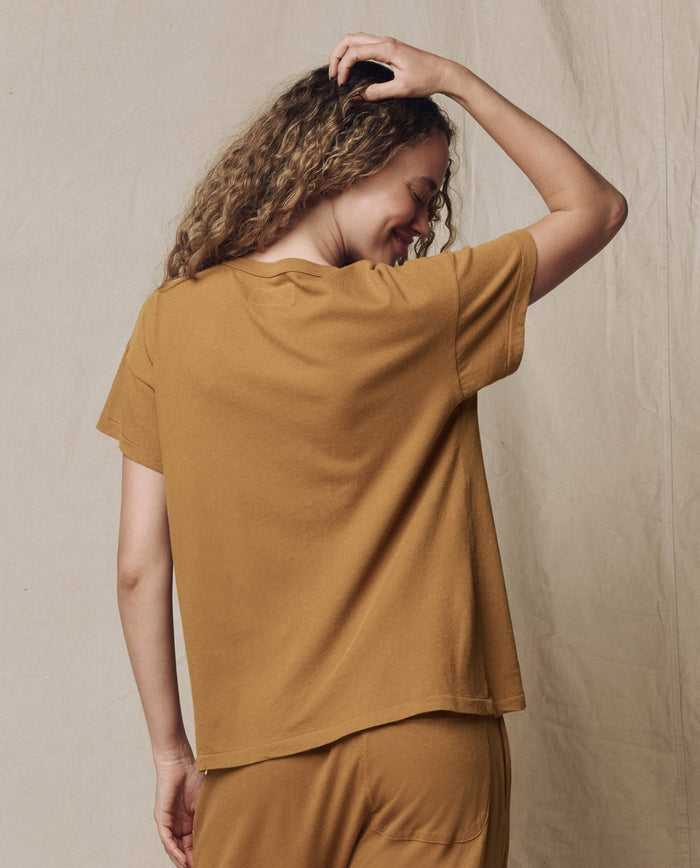 A person with curly hair wears THE GREAT. SU26 SEASONAL CORE The Boxy Crew. Solid in Cork, paired with matching pants, standing facing away from the camera with one arm raised against a plain beige background.