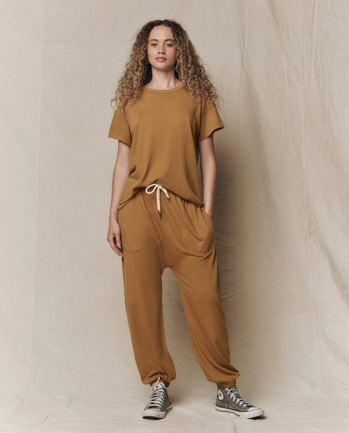 A person stands against a neutral backdrop, wearing THE GREAT.'s The Boxy Crew. Solid in Cork with matching drawstring pants featuring pockets, paired with gray high-top sneakers.