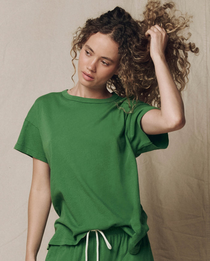 A woman wears THE GREAT.'s The Boxy Crew in Solid Fennel (SU26 SEASONAL CORE) with matching shorts, standing against a neutral background and holding up her curly hair as she looks to the side.