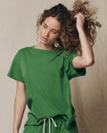 A woman wears THE GREAT.'s The Boxy Crew in Solid Fennel (SU26 SEASONAL CORE) with matching shorts, standing against a neutral background and holding up her curly hair as she looks to the side.
