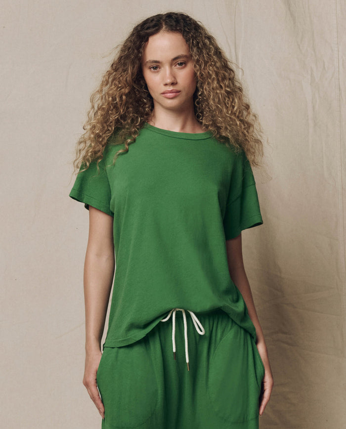 A woman with curly hair wears THE GREAT.'s The Boxy Crew in Solid Fennel, paired with matching green drawstring pants, standing against a neutral background.