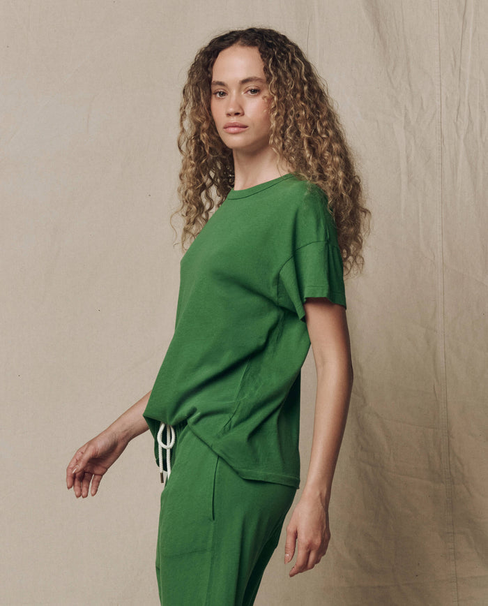 A person with long curly hair wears THE GREAT. SU26 SEASONAL CORE The Boxy Crew. Solid -- Fennel t-shirt and matching green pants, standing before a beige fabric backdrop.