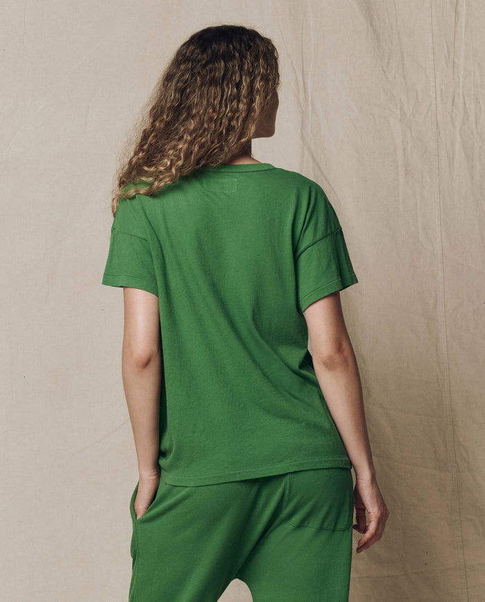 A person with curly hair stands with their back to the camera, wearing THE GREAT.'s Boxy Crew Solid t-shirt in Fennel from the SU26 SEASONAL CORE collection, paired with green pants against a plain, light-colored background.
