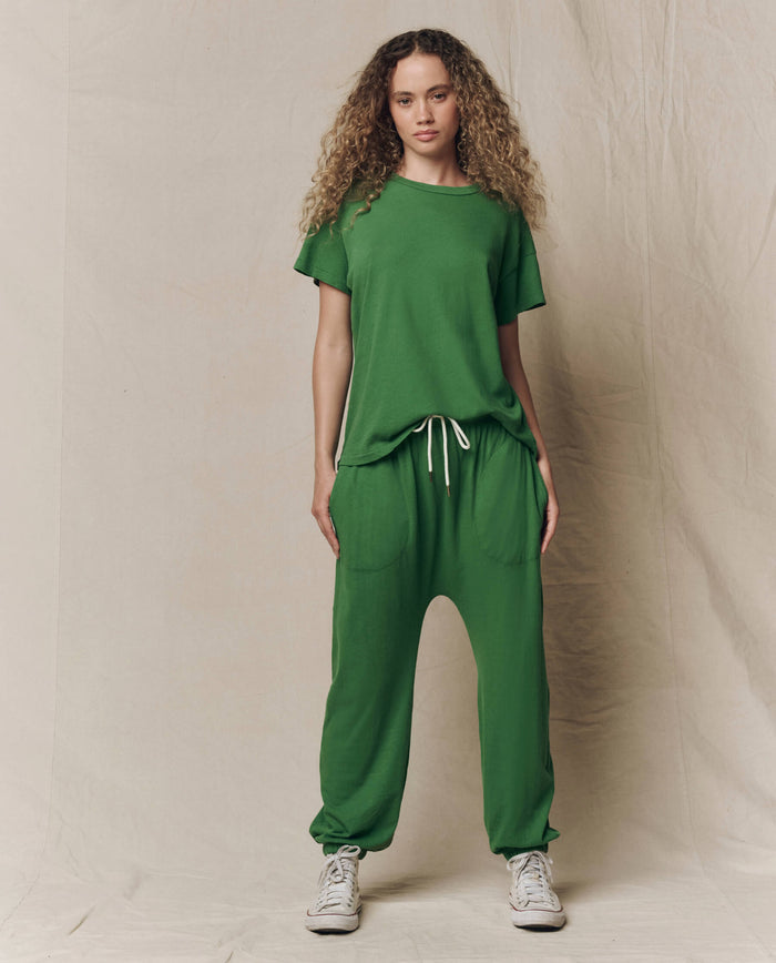 A person stands facing forward wearing THE GREAT. SU26 SEASONAL CORE The Boxy Crew. Solid -- Fennel, paired with matching green drawstring sweatpants and white sneakers, set against a plain beige backdrop.