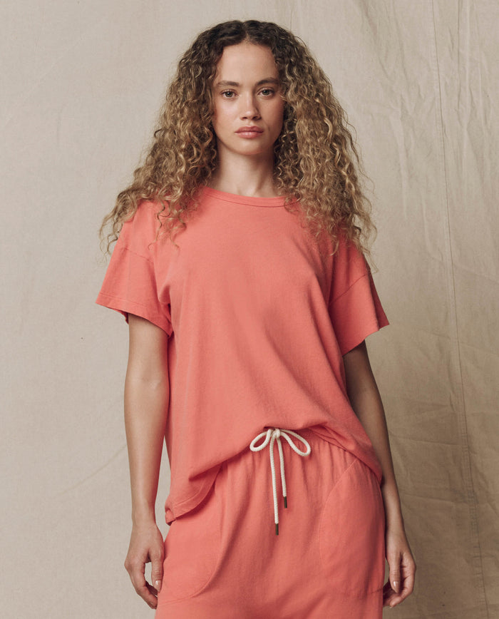 A person with long curly hair wears THE GREAT.’s The Boxy Crew. Solid -- Guava and matching pants with a white drawstring, standing against a neutral backdrop.