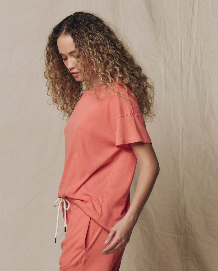 A person with curly hair wears THE GREAT. SU26 SEASONAL CORE The Boxy Crew. Solid -- Guava, standing in profile against a neutral fabric backdrop.