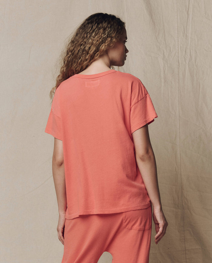 Person with long curly hair, seen from behind, wears THE GREAT. SU26 SEASONAL CORE The Boxy Crew. Solid in Guava with matching pants, standing against a neutral fabric backdrop.