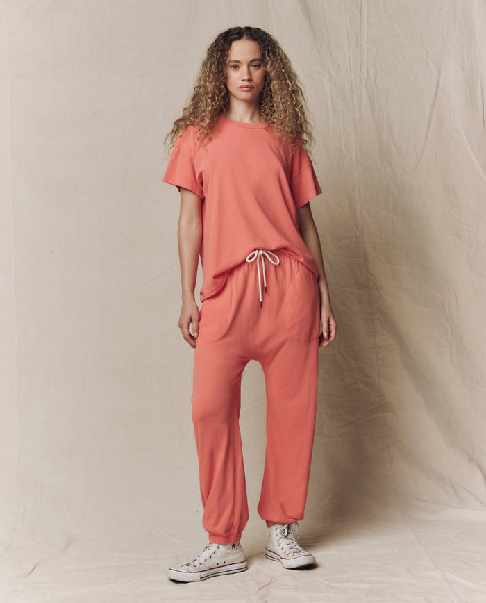 A woman stands against a beige fabric backdrop wearing THE GREAT.'s The Boxy Crew. Solid in Guava from the SU26 SEASONAL CORE collection, paired with matching drawstring pants and white sneakers.