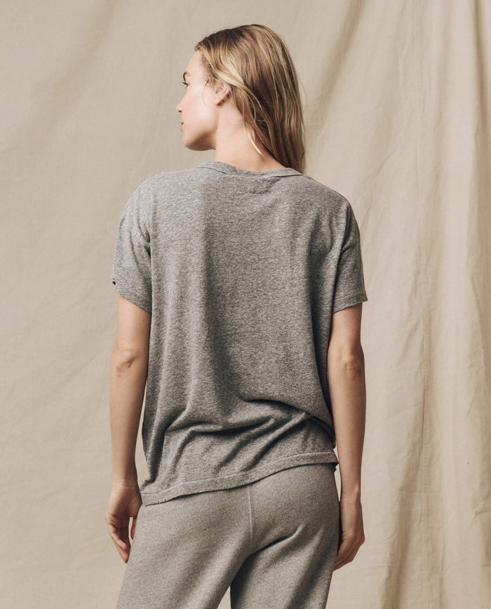 A person with long hair stands facing away, showcasing a loose-fitting vintage tee and gray pants against a beige backdrop. The Boxy Crew in Heather Grey from THE GREAT. CORE KNITS, made in the U.S.A., features flattering tweaks that set it apart.