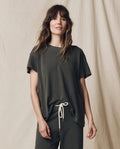 A person with long hair stands against a neutral backdrop, wearing THE GREAT. CORE KNITS' The Boxy Crew in Solid -- Washed Black, paired with drawstring pants. Made from 100% cotton in the U.S.A., this outfit exudes a classic vibe reminiscent of vintage tees.